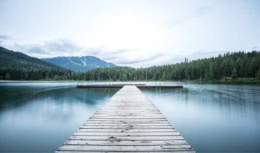 A photo of a dock on a lake, with trees and mountains in the background.