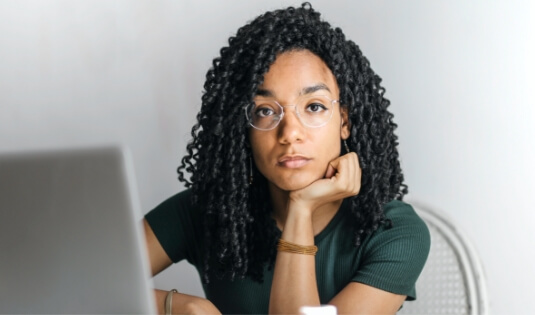 multi-racial woman sits in front of a laptop, with her chin resting on her hand.